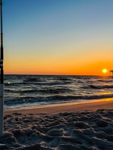 Fishing rod in the sand at sunset next to a pier with people on it